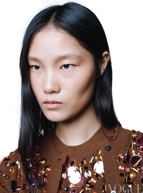 Portrait of an Asian female model with dark hair and a brown embellished cardigan, posing in a studio with a white background.