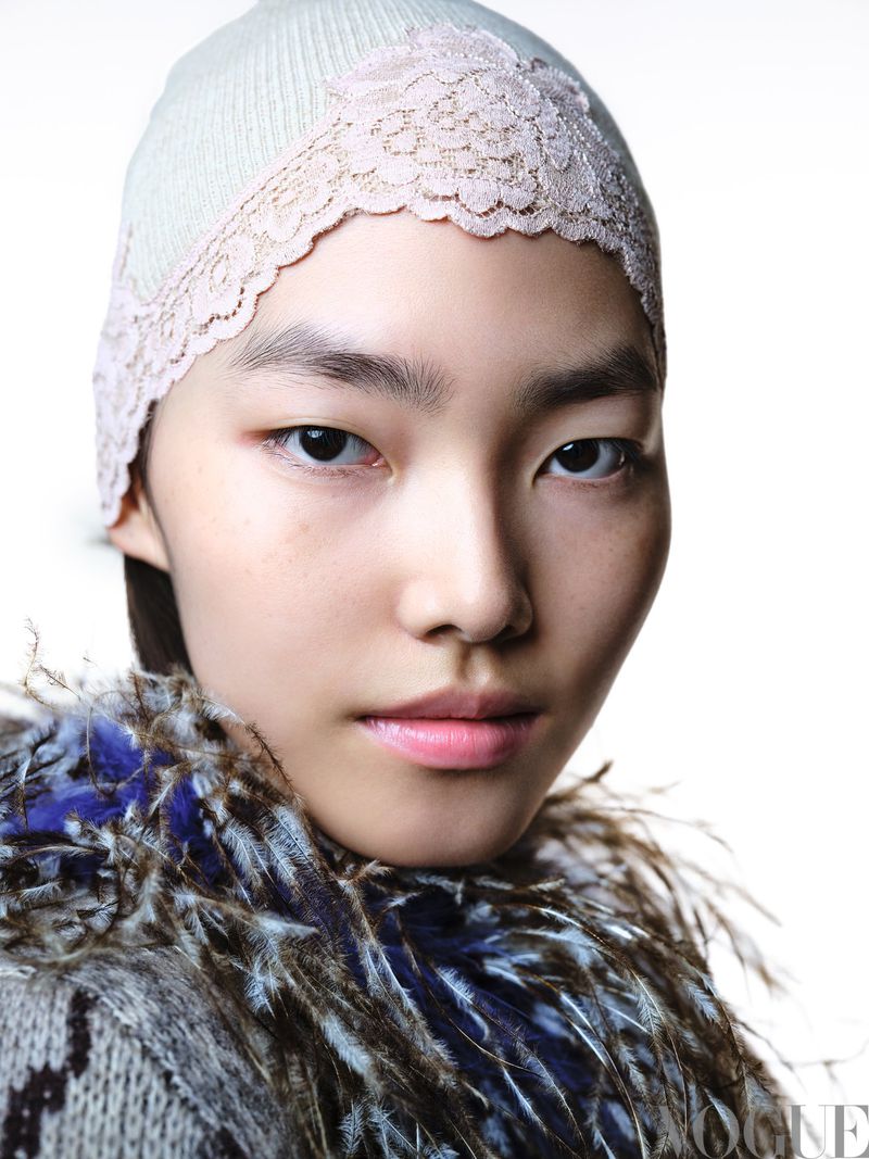 Close-up portrait of a young Asian woman wearing a knitted hat with lace trim and a feathered collar against a bright white background.