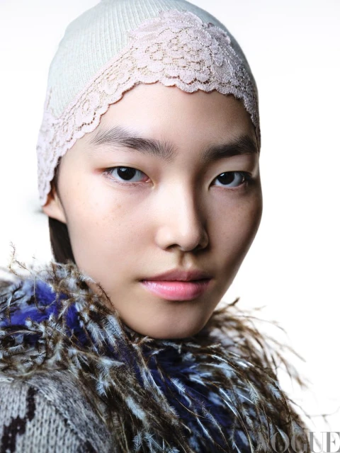 Close-up portrait of a young Asian woman wearing a knitted hat with lace trim and a feathered collar against a bright white background.