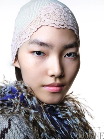Close-up portrait of a young Asian woman wearing a knitted hat with lace trim and a feathered collar against a bright white background.