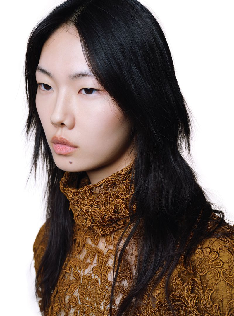 Close-up studio shot of a beautiful Asian female fashion model with black hair wearing a brown lace turtleneck top against a white background.