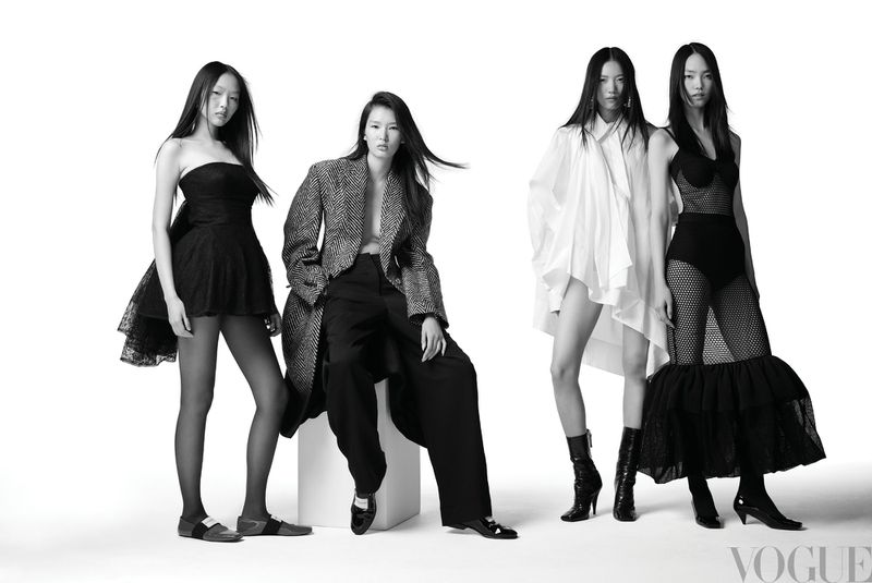 Four East Asian women model black and white fashion outfits in a bright, high-key studio shot for Vogue. Full body view, stark lighting.