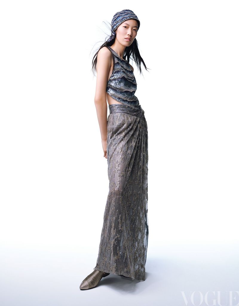 A fashionable Asian woman poses in a sleeveless silver top and matching skirt and headwrap, set against a bright white backdrop.