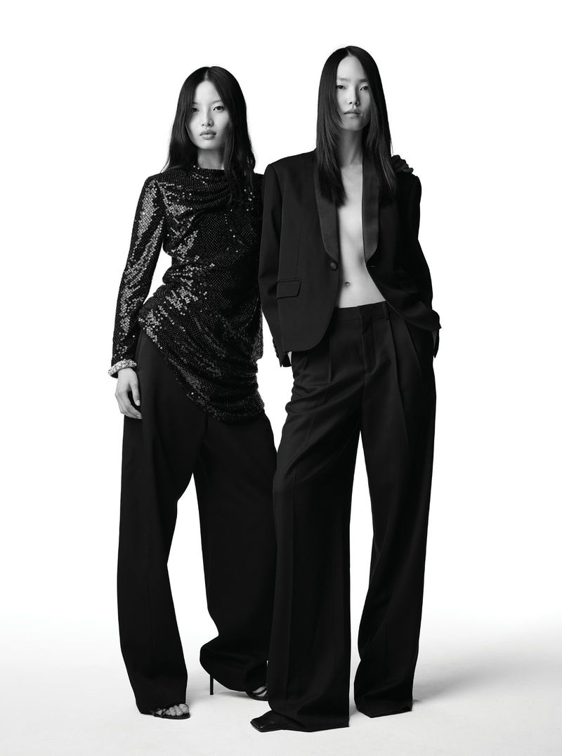 Two Asian women in black evening wear stand in a bright studio. One wears a sequin top and the other a blazer with wide-leg pants.