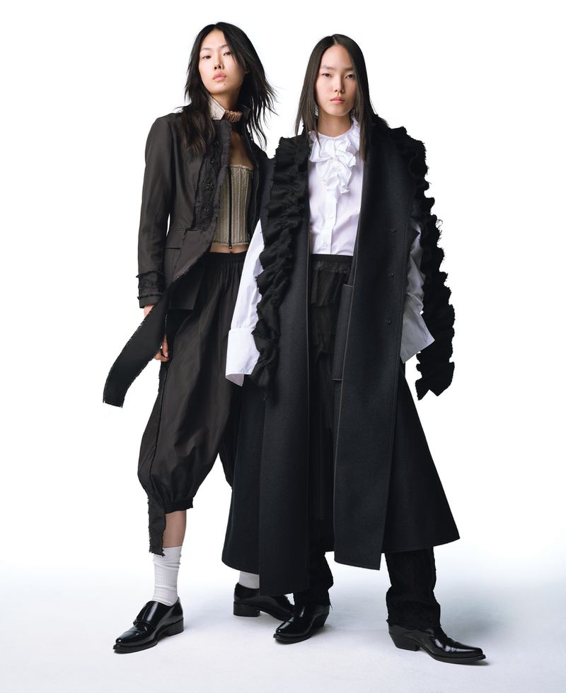 Two Asian fashion models in black and white avant-garde outfits pose in a studio, showcasing layered tops, wide-leg pants, and boots on a white backdrop.