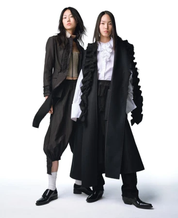 Two Asian fashion models in black and white avant-garde outfits pose in a studio, showcasing layered tops, wide-leg pants, and boots on a white backdrop.