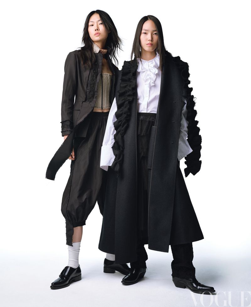 Two Asian fashion models in dark, avant-garde garments pose against a stark white background in a studio, with Vogue logo present.