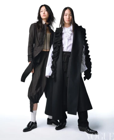 Two Asian fashion models in dark, avant-garde garments pose against a stark white background in a studio, with Vogue logo present.