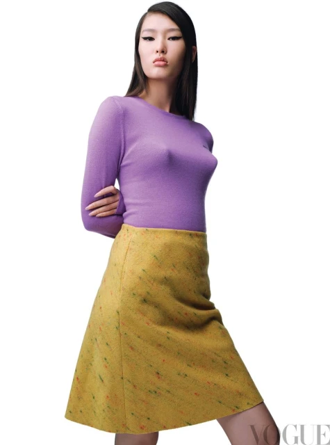 Asian woman in a studio setting is wearing a purple top and patterned yellow skirt, posing for a fashion shot with a white backdrop.