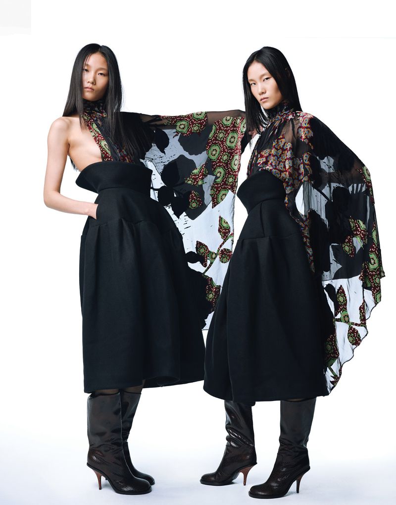Two Asian women models in high-fashion attire pose against a white backdrop, wearing dramatic black skirts, sheer patterned tops and boots.