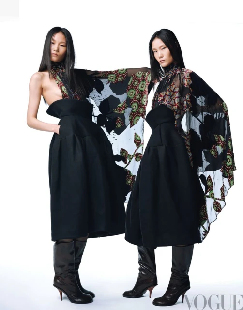 Two Asian female models in fashionable black dresses and boots pose in a studio, showcasing unique cape-like garments, fashion shoot.