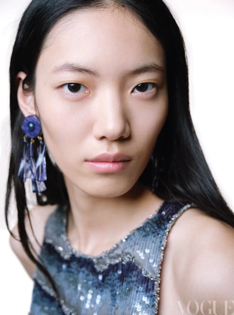 Close-up fashion portrait of an Asian adult woman with dark hair wearing a blue sequin top and blue floral earrings in a bright studio.
