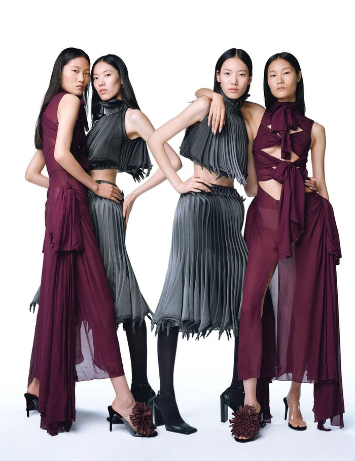 Four female models posing in pleated grey and burgundy outfits against a solid white background