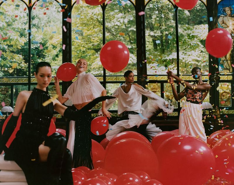 Group of four adult female fashion models in elegant gowns are surrounded by a field of red balloons and confetti in a greenhouse.