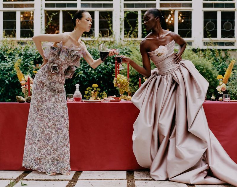 Two women in elegant gowns raise wine glasses at an outdoor event with flowers, candles, and a red tablecloth in a garden setting.