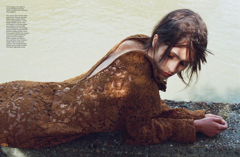 Woman in brown lace dress lying on stone near water, posing with eyes closed in a fashion editorial portrait with soft lighting.