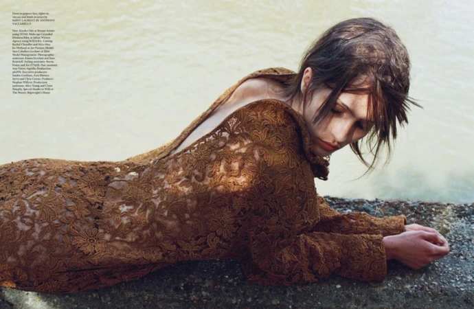 Woman in brown lace dress lying on stone near water, posing with eyes closed in a fashion editorial portrait with soft lighting.