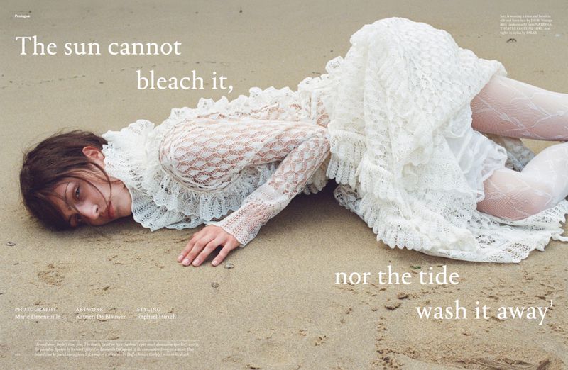 Woman in a white lace dress lies on a sandy beach, looking at the camera with a neutral expression in a fashion editorial.