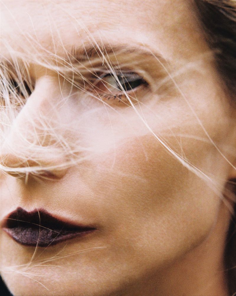 Close-up portrait of a woman with light hair partially covering her face, dark lipstick, and fair skin, highlighting beauty and fashion.