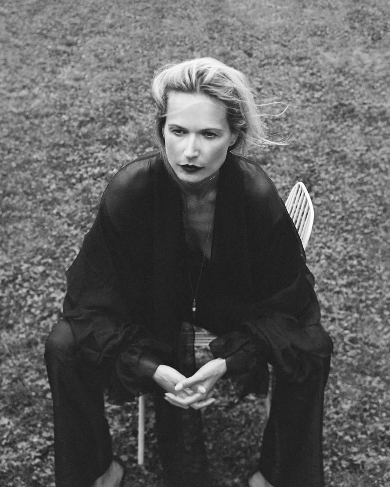 Black and white portrait of a woman sitting in a chair outdoors, wearing a dark outfit, with short blond hair and dark lipstick.