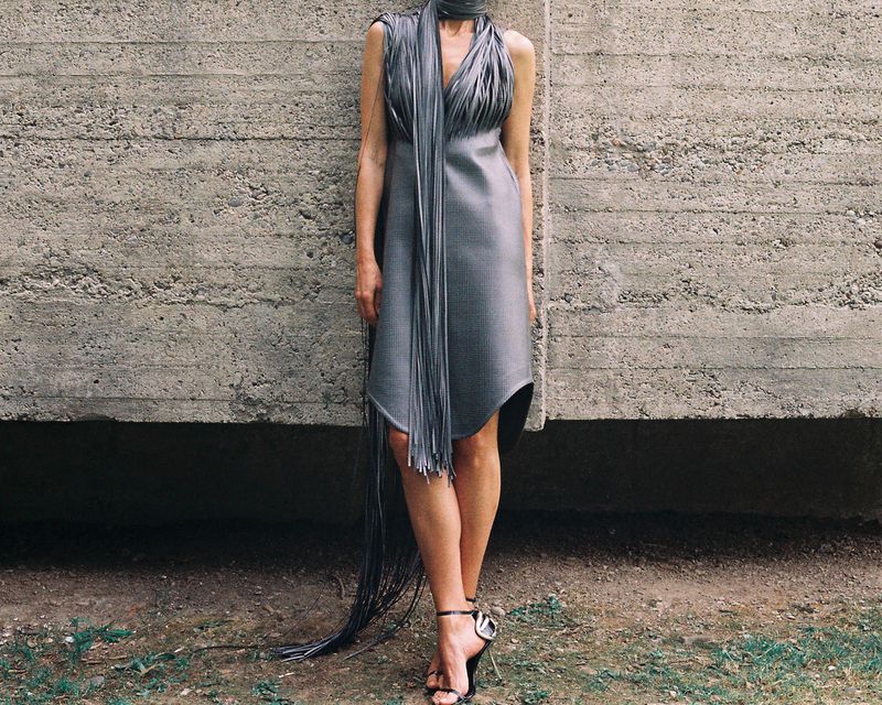 A woman in a grey dress with long fringes stands against a concrete wall in natural light, wearing high-heeled sandals.