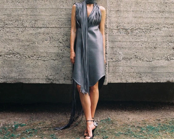 A woman in a grey dress with long fringes stands against a concrete wall in natural light, wearing high-heeled sandals.