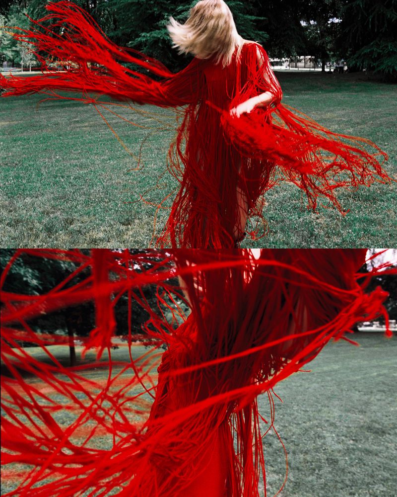 Adult female wearing a red fringed dress dances in a grassy field surrounded by trees, with a blurred motion effect highlighting movement.