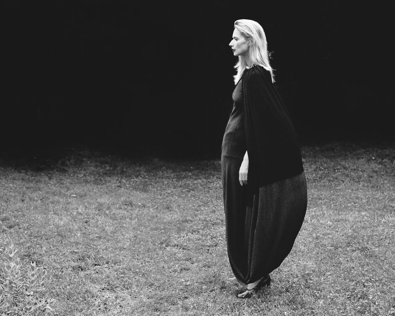 Black-and-white profile shot of a blonde woman in elegant flowing pants and a draped top posing in a grassy outdoor setting against a dark backdrop.
