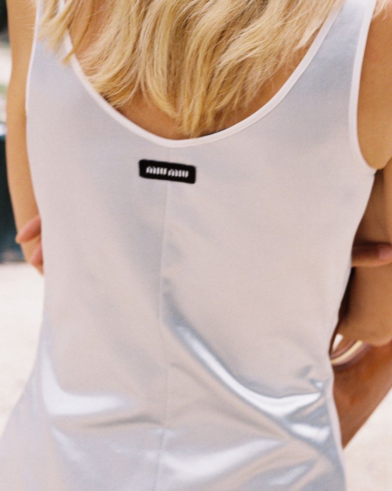 Blonde woman in a white, metallic-look dress, back to the camera, featuring a Miu Miu logo tag, shot in bright, natural outdoor lighting.