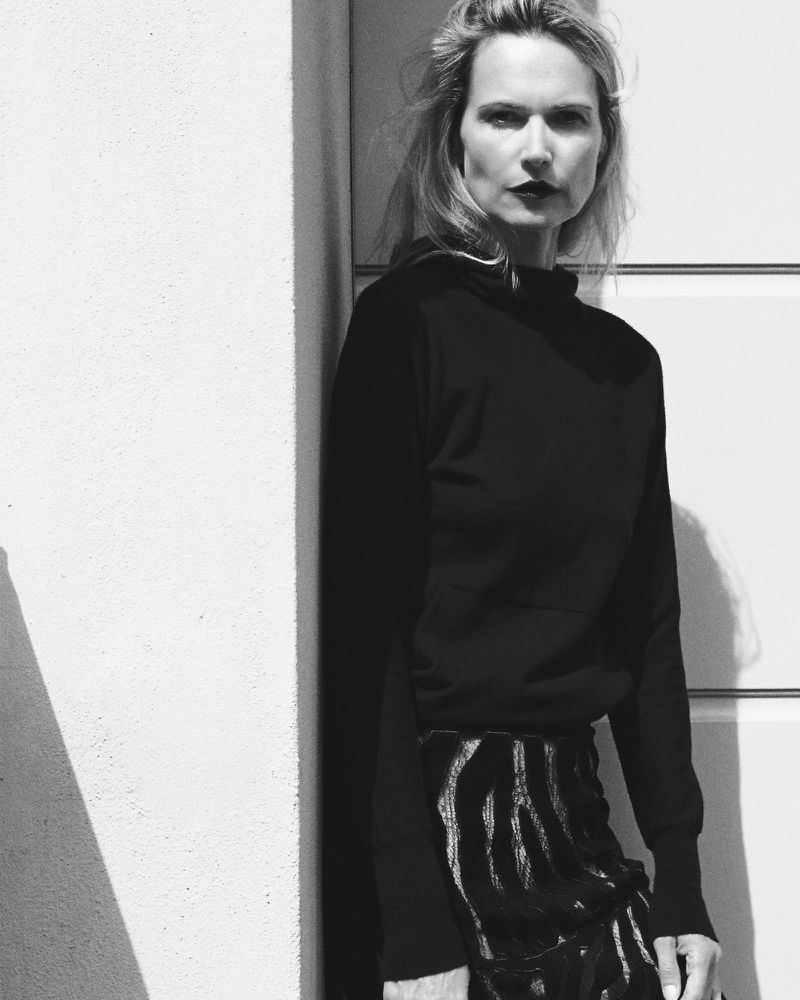Monochrome portrait of a stylish woman in a black top and patterned skirt posing against a white wall with geometric shadows.