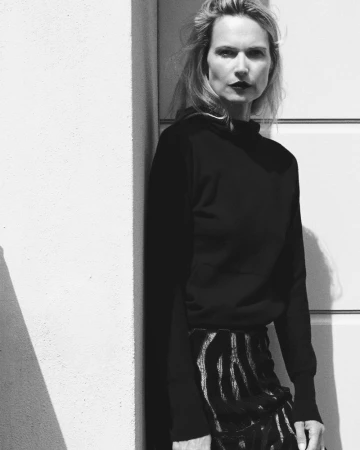 Monochrome portrait of a stylish woman in a black top and patterned skirt posing against a white wall with geometric shadows.