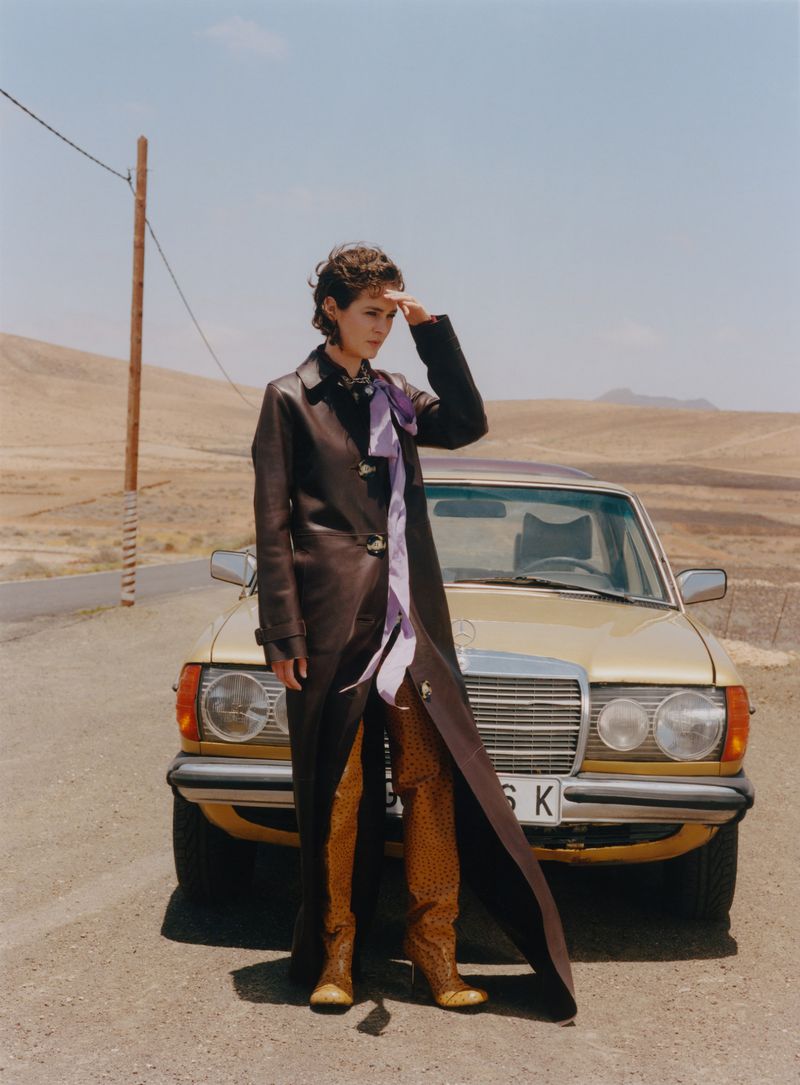 A woman in a long brown leather coat shields her eyes next to a gold Mercedes-Benz car on a desert road under a blue sky.