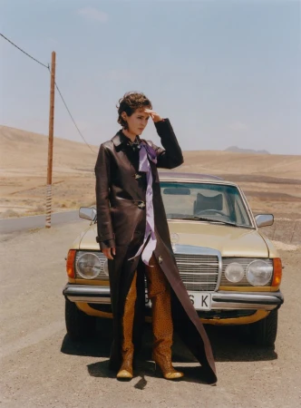 A woman in a long brown leather coat shields her eyes next to a gold Mercedes-Benz car on a desert road under a blue sky.