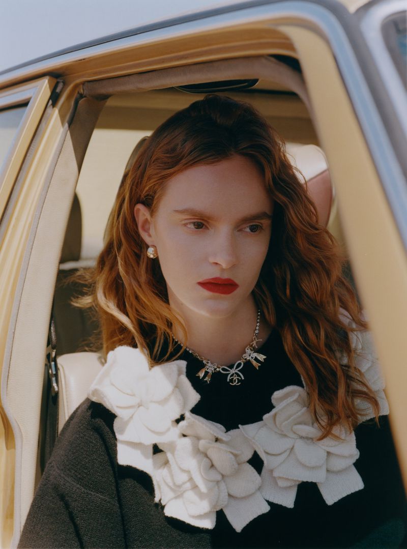 Portrait of a red-haired woman with fair skin in the passenger seat of a car wearing a Chanel necklace and sweater.