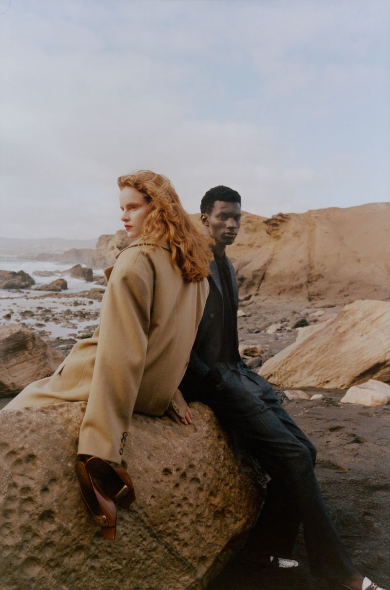 Two models, a redhead woman and a black man, pose on a large rock at the beach with a cloudy sky background and sea.