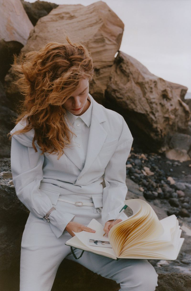 Woman with red hair sitting on rocky beach reading a notebook wearing white blazer and pants against rock formations and neutral sky.