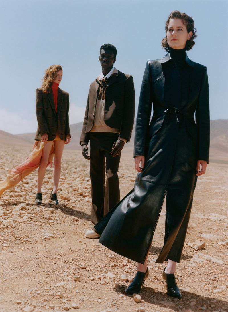 Three fashion models in an outdoor desert setting wear blazers and pantsuits. A red scarf and bright blue sky add vibrant color.
