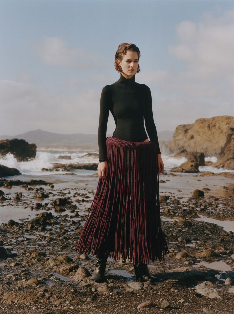 A woman stands on a rocky beach wearing a black turtleneck and a long, fringed maroon skirt with the ocean and sky in the background.