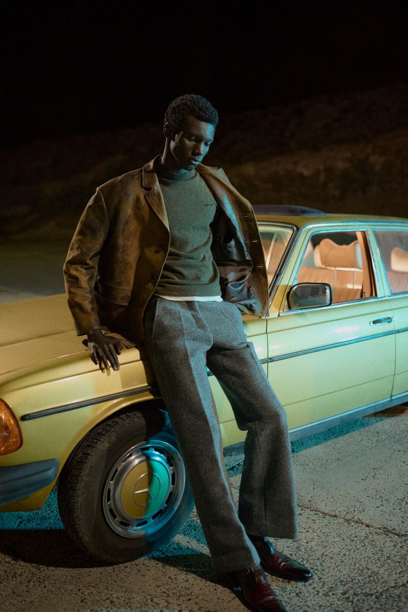 An African-American man leans on a yellow vintage car at night, wearing a brown jacket, green sweater, grey pants and burgundy shoes.