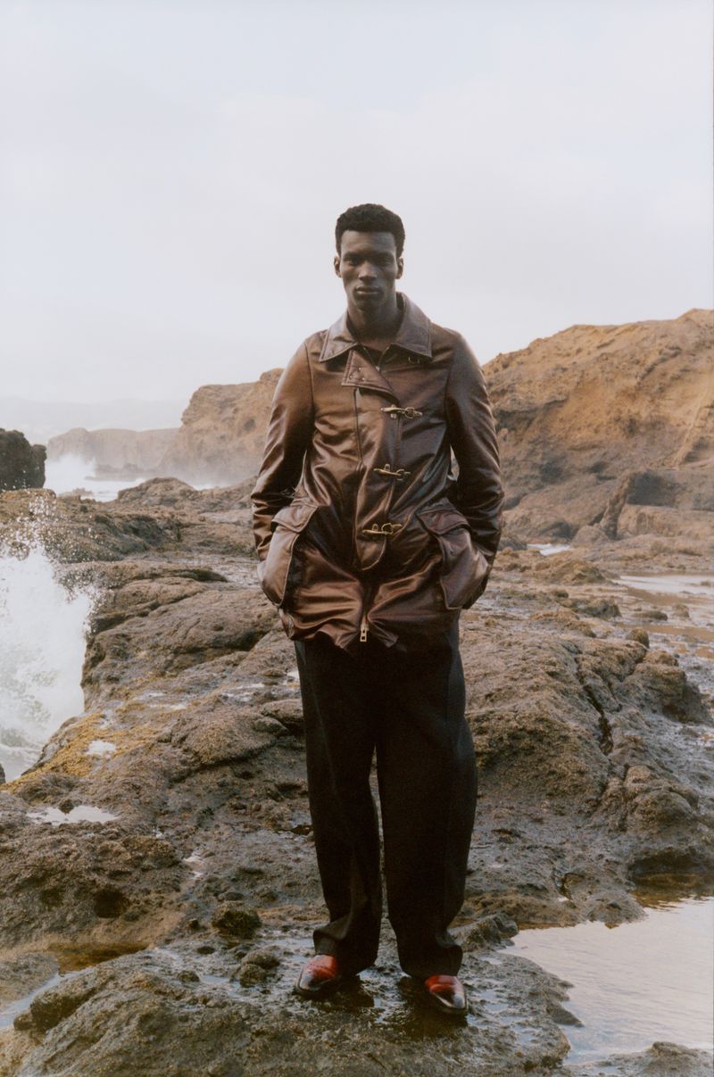 A dark-skinned male model poses on a rocky beach in a brown jacket and black trousers, near ocean waves under an overcast sky.