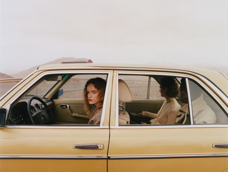 Two women inside a classic yellow Mercedes-Benz in an arid landscape, one at the wheel with red lipstick.