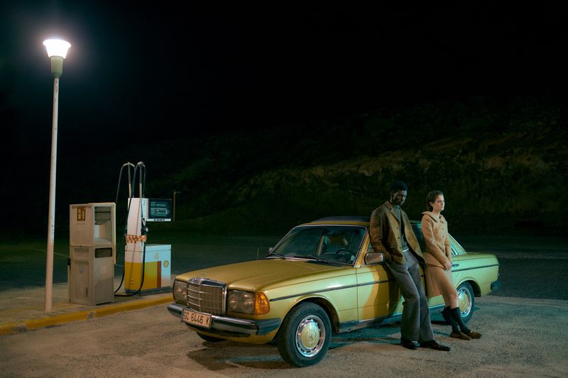 An interracial couple stands by a classic yellow car at night, parked beside a vintage gas station under the glow of a street lamp.