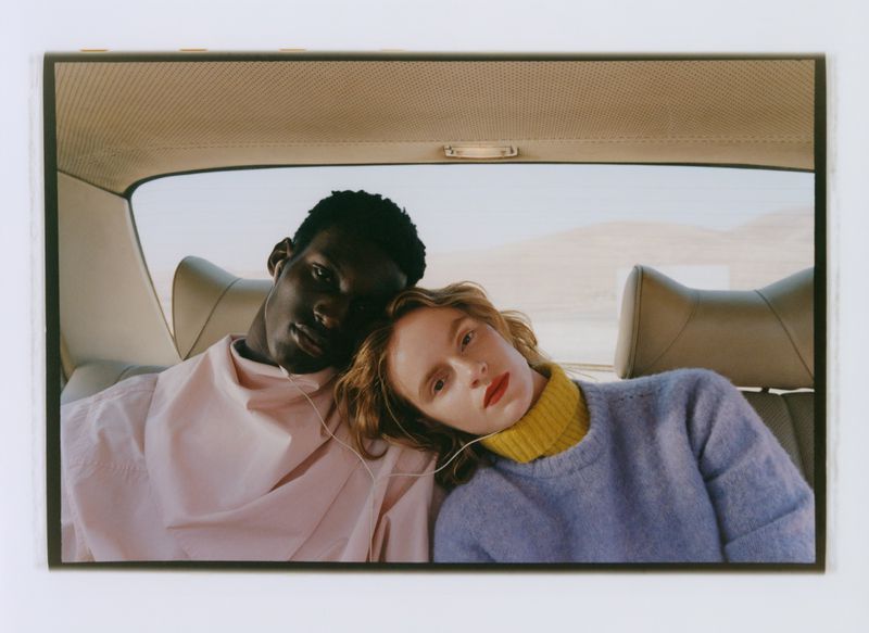 A young interracial couple in a car interior. The woman is leaning on the man's shoulder, both looking towards the camera.