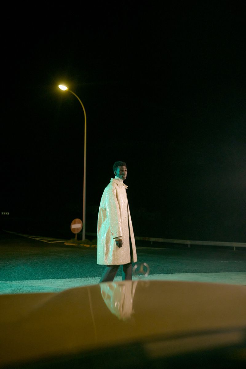 An adult man wearing a light tan coat standing on an outdoor road illuminated by a street light against a dark night sky.