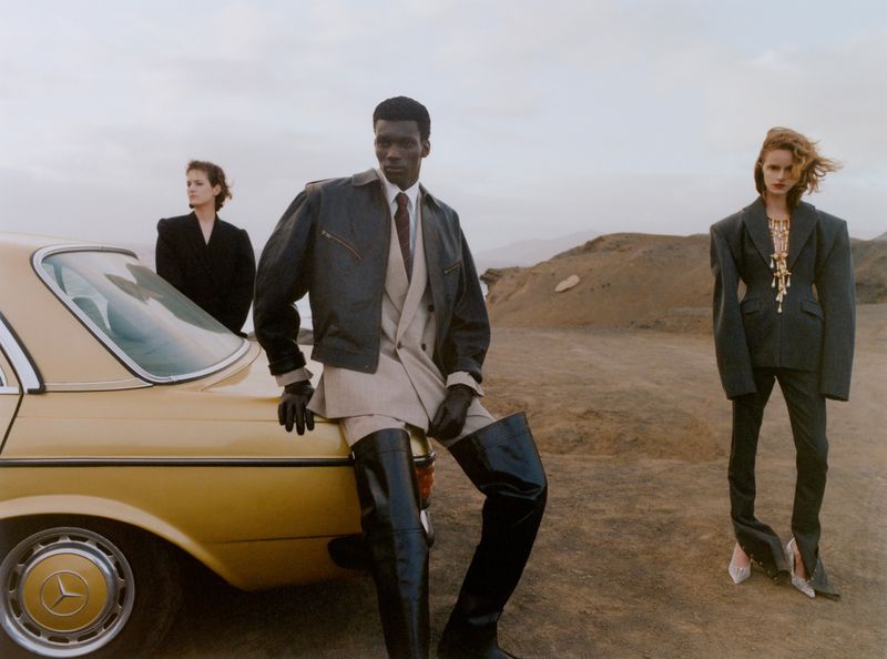 Fashion models pose beside a vintage car in a desert landscape, wearing structured suits and boots, exuding a blend of retro and modern style.