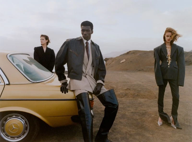 Fashion models pose beside a vintage car in a desert landscape, wearing structured suits and boots, exuding a blend of retro and modern style.