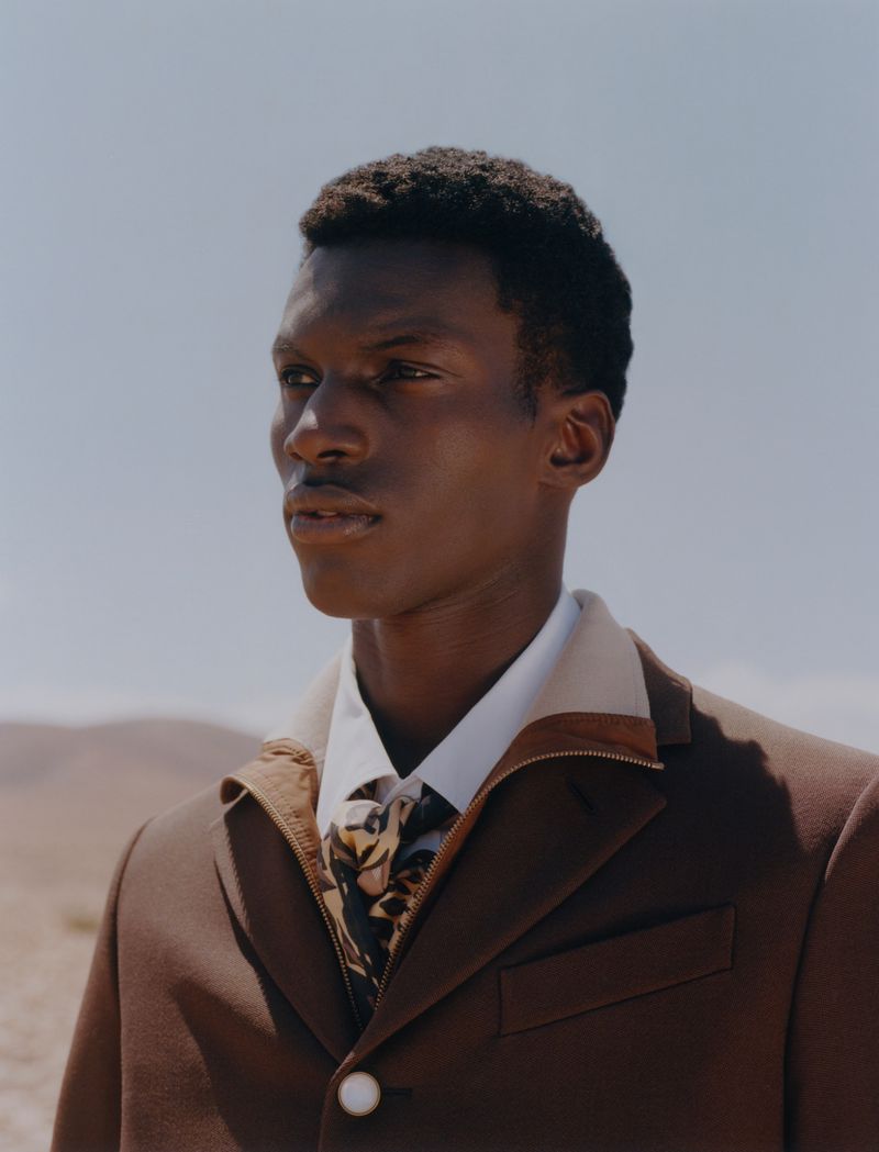 A dark-skinned male model poses in a brown jacket, white collared shirt, and tie in an outdoor desert landscape with a blue sky.