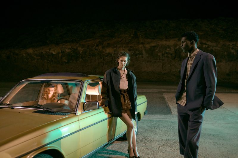 A vintage yellow car sits with two women inside, while a woman leans on the door and a man stands to the side in an outdoor night scene.