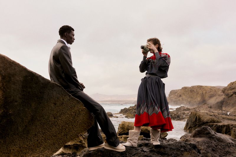A man in a suit sitting on a rock looking away while a woman stands opposite him taking a photograph with a vintage camera.