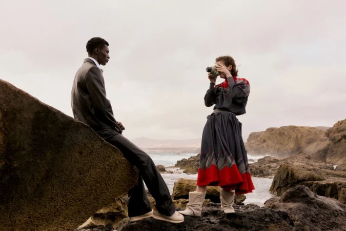 A man in a suit sitting on a rock looking away while a woman stands opposite him taking a photograph with a vintage camera.